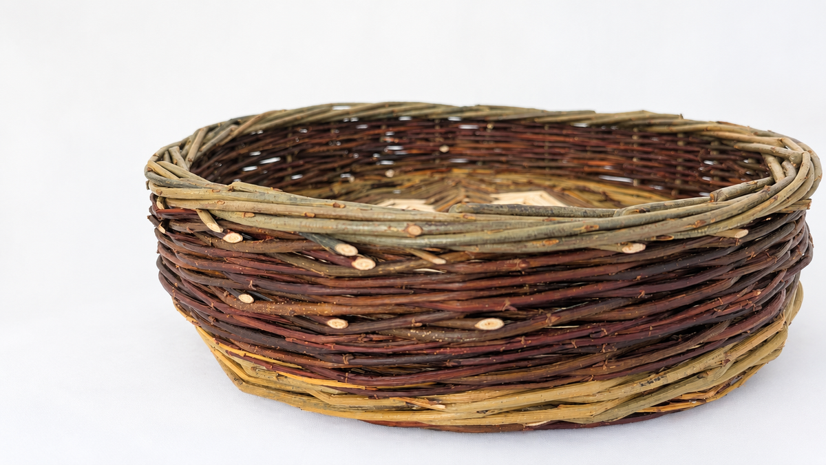 A round woven basket made from natural willow branches, featuring a rustic design with varying shades of brown, tan, and olive green, set against a plain white background.